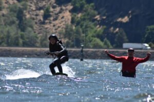 Two wakeboarders ride a lake, one in black gear and helmet on a green board, the other in red shirt, both arms raised in excitement with a hillside backdrop.