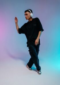 Young man dancing in an oversized black shirt and dark jeans, wearing white headphones, against a pastel gradient backdrop. It captures a mid-step pose with one hand raised.