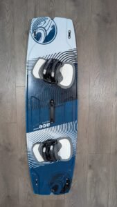 Blue and white wakeboard with black bindings mounted, lying on a wooden floor with circular graphic patterns on the board edges.