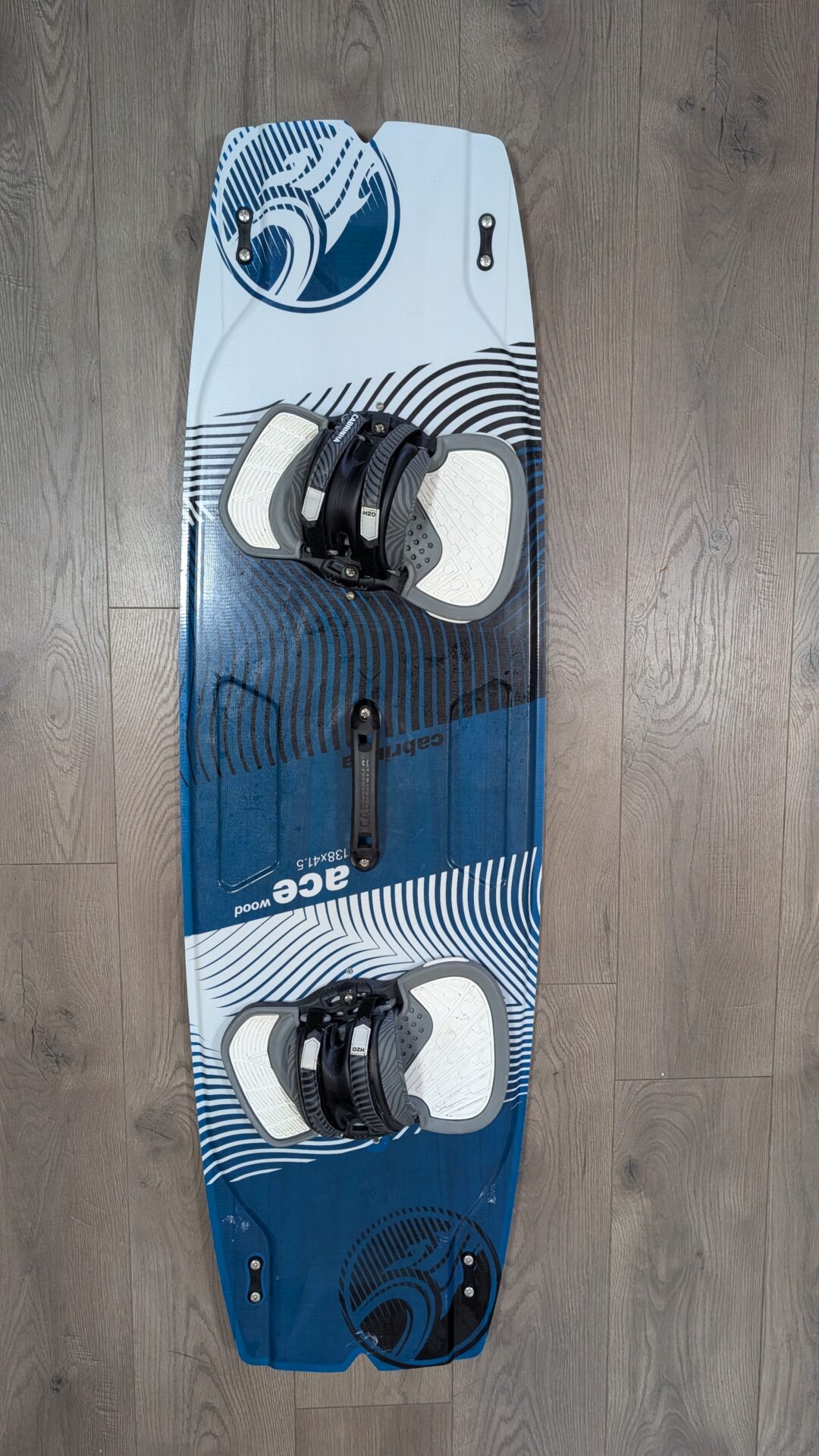 Blue and white wakeboard with black bindings mounted, lying on a wooden floor with circular graphic patterns on the board edges.