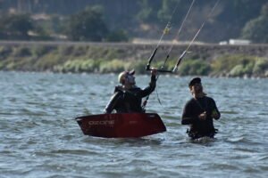 Two men in wetsuits with a red Ozone kiteboard boat in the water, handling kite control bars as lines rise overhead.