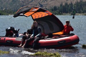 Three people in wetsuits sit on a red inflatable boat with a large orange and black kite overhead on a lake, mountains in the background.