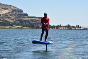 Stand-up paddler wearing a red long-sleeve shirt and cap on a blue SUP, paddling on a lake with rocky hills in the background on a sunny day.