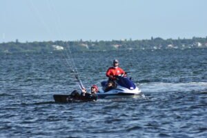 Man in a red shirt on a blue and white jet ski, with another person in a small boat nearby on a windy sea; coastline in the distance.