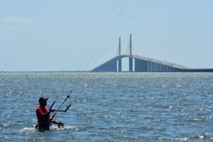 Kiteboarder in a red shirt and sunglasses stands in shallow water, holding the control bar as lines rise overhead, with a long bridge in the background of the sea.