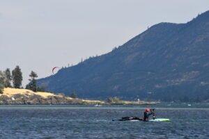 Kite surfer with a red arc-shaped kite on a lake with mountains in the background and a rocky shoreline in the foreground