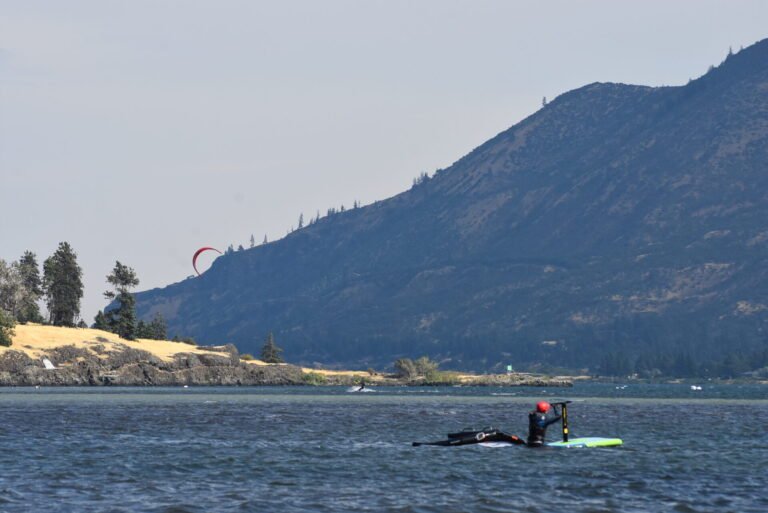 Kite surfer with a red arc-shaped kite on a lake with mountains in the background and a rocky shoreline in the foreground