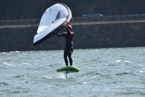 Windsurfer wearing a pink helmet and wetsuit on a foil board, handling a large white sail on choppy water outdoors.