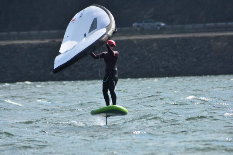 Windsurfer wearing a pink helmet and wetsuit on a foil board, handling a large white sail on choppy water outdoors.