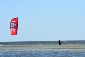 Kite surfer riding on choppy ocean water with a large red kite flying on the left under a clear blue sky.