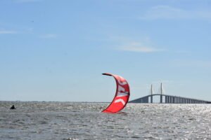 Kite surfer riding choppy water near a long bridge, with a red curved kite in the foreground.