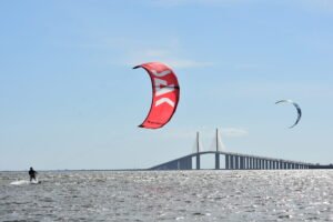 Kite surfer riding on choppy water near a large bridge, with a bright red kite with white lettering in the foreground.