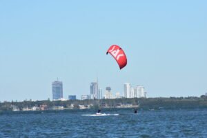 Kite surfer on a red kite skimming over dark blue water with a city skyline in the distance under a clear sky.