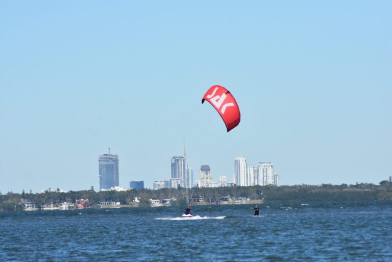 Kiteboard Lessons in St Petersburg Fl - Tampa Bay 46 Kite surfer on a red kite skimming over dark blue water with a city skyline in the distance under a clear sky.