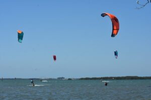 Kitesurfer on the left riding in choppy water under a clear blue sky, colorful kites in the air above.