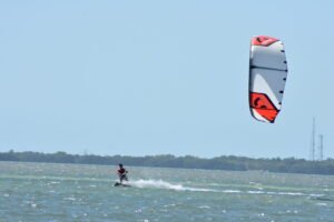 Kiteboarder riding on blue sea with a large red-and-white kite in clear sky.