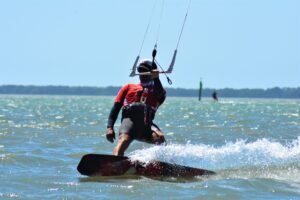Kiteboarder wearing a red shirt, helmet and sunglasses rides a board over choppy water, holding the bar in bright sunlight.