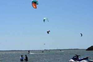 Kiteboarders glide on choppy water under a clear blue sky, with colorful kites high above the horizon and a jet ski in the foreground.