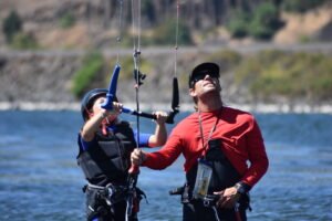 Two people in life vests hold kite lines and a control bar in shallow water, preparing to kiteboard. (informative)
