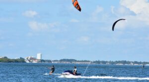 Two kiteboarders ride the choppy sea while a jet ski follows, with a distant city coastline and buildings on the horizon.