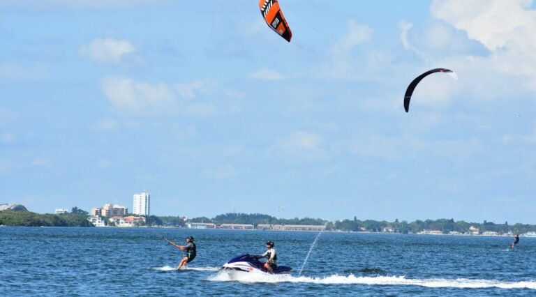 Kiteboard Lessons in St Petersburg Fl - Tampa Bay 45 Two kiteboarders ride the choppy sea while a jet ski follows, with a distant city coastline and buildings on the horizon.