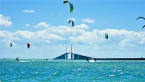 Kite surfers riding choppy turquoise water under a blue sky with a distant bridge in the background