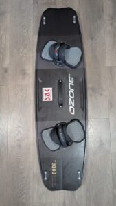 Black wakeboard with gray bindings attached, lying on a wood floor; shows Ozone branding and a red accent on the binding strap packet center area.
