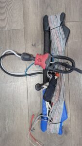 Kite control bar with colorful lines coiled around, resting on a wooden floor, ready to be used.