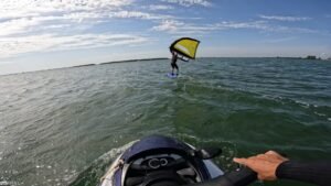 Person windsurfing on a blue board with a yellow sail, gliding across calm water under a cloudy sky, seen from a nearby craft
