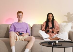 Two teens sit on a beige sofa playing a console game with controllers, pink and yellow ambient lighting in the background.