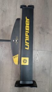 Carbon fiber foil wing placed next to a tall black case with yellow branding on a wooden floor.